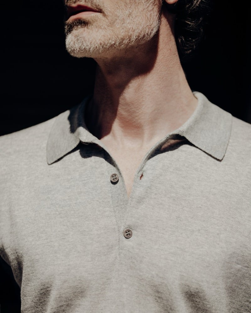Tight chest-up crop of Richard Biedul in the John Smedley Isis polo in heather gray, showing the fine-gauge Sea Island Cotton knit, soft collar, and two-button placket in raking sunlight