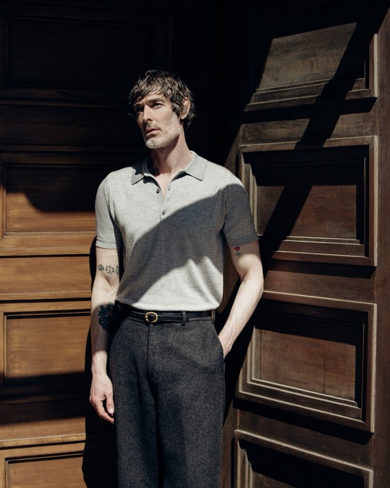 Richard Biedul standing in a doorway of dark wood paneling in the John Smedley Isis knitted polo in pale heather gray, tucked into charcoal pleated wool trousers held by a thin black leather belt with a small gold buckle