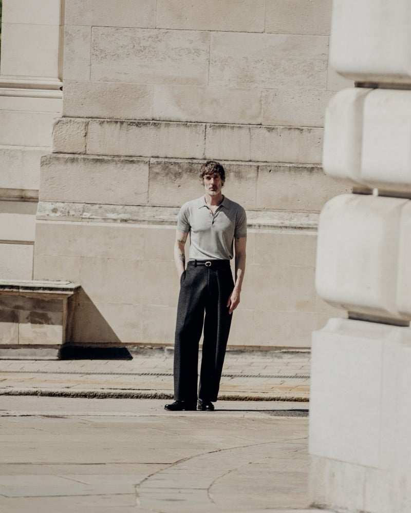 Full-length frame of Richard Biedul standing in a stone courtyard of a London neoclassical building in the gray John Smedley Isis polo tucked into charcoal pleated wide-leg trousers with a thin belt and black loafers
