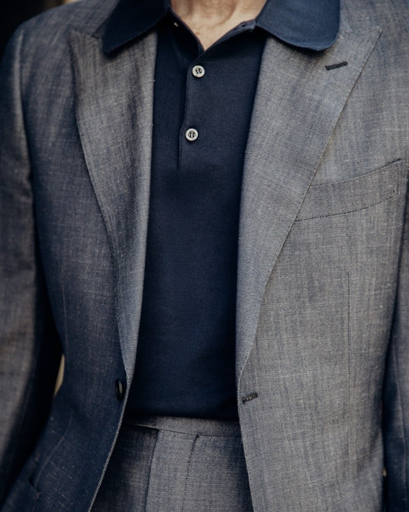 Detail crop of the navy John Smedley Oakman polo worn by Richard Biedul under an open blue-gray melange wool jacket, focused on the three-button placket and soft knitted collar
