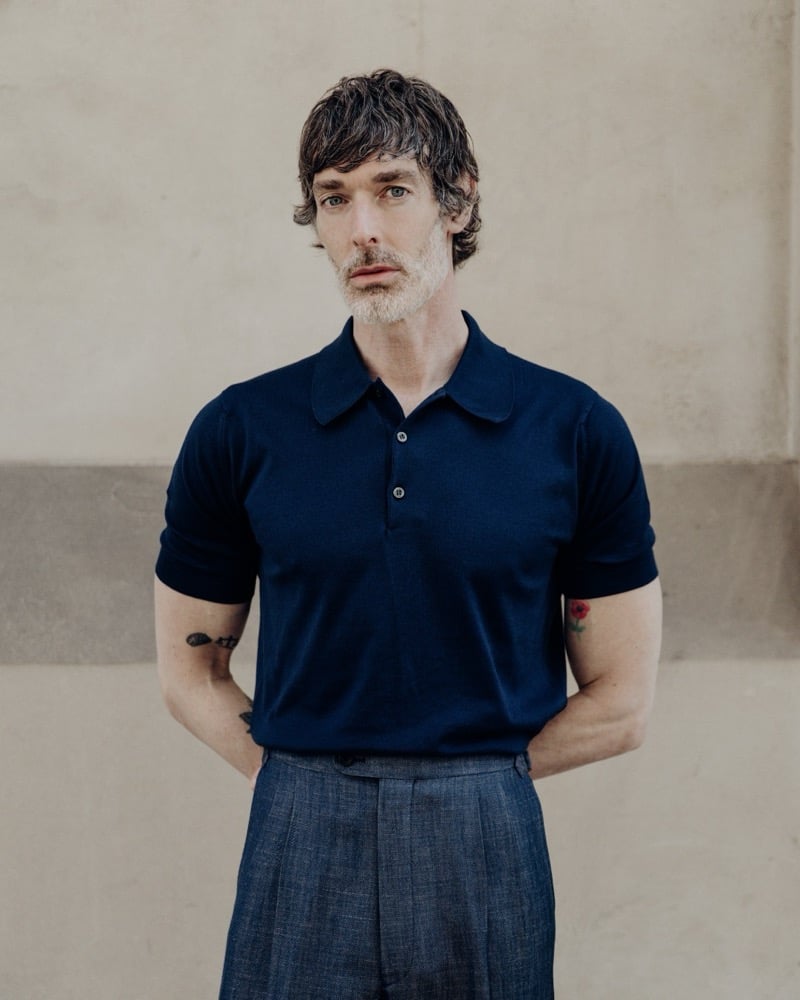 Mid-body portrait of Richard Biedul against a pale stone wall wearing the John Smedley Oakman knitted polo in navy with a relaxed easy fit, tucked into slubby mid-blue pleated wide-leg trousers
