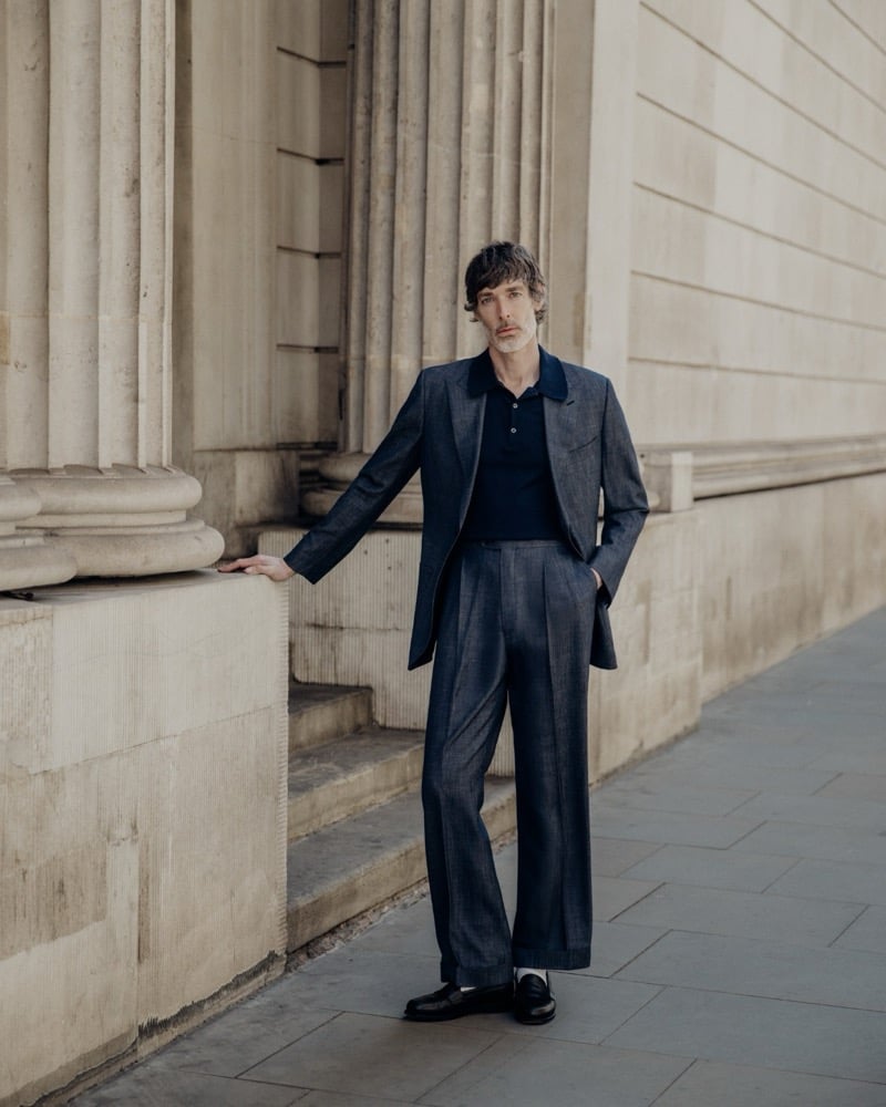 Richard Biedul stands beside a column of a London neoclassical building in a John Smedley Oakman knitted polo worn under an open blue-gray melange wool jacket with matching pleated wide-leg trousers and black leather loafers