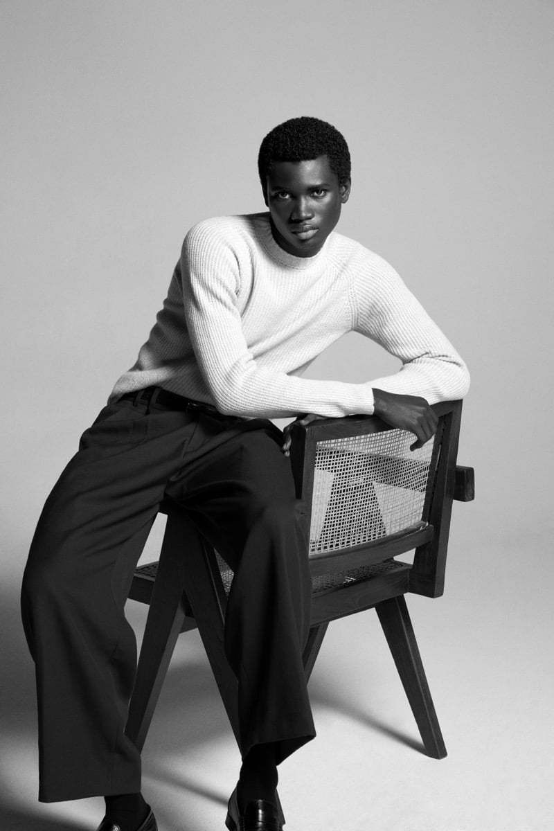 Alexander Acquah leans against a Pierre Jeanneret-style chair in the John Smedley Oxnard eco cashmere ribbed crewneck
