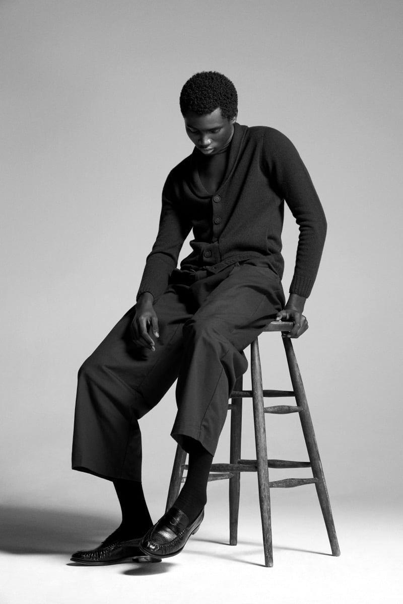 Alexander Acquah perches on a wood stool in the John Smedley Rockford eco cashmere shawl-collar cardigan layered over a turtleneck with cropped trousers
