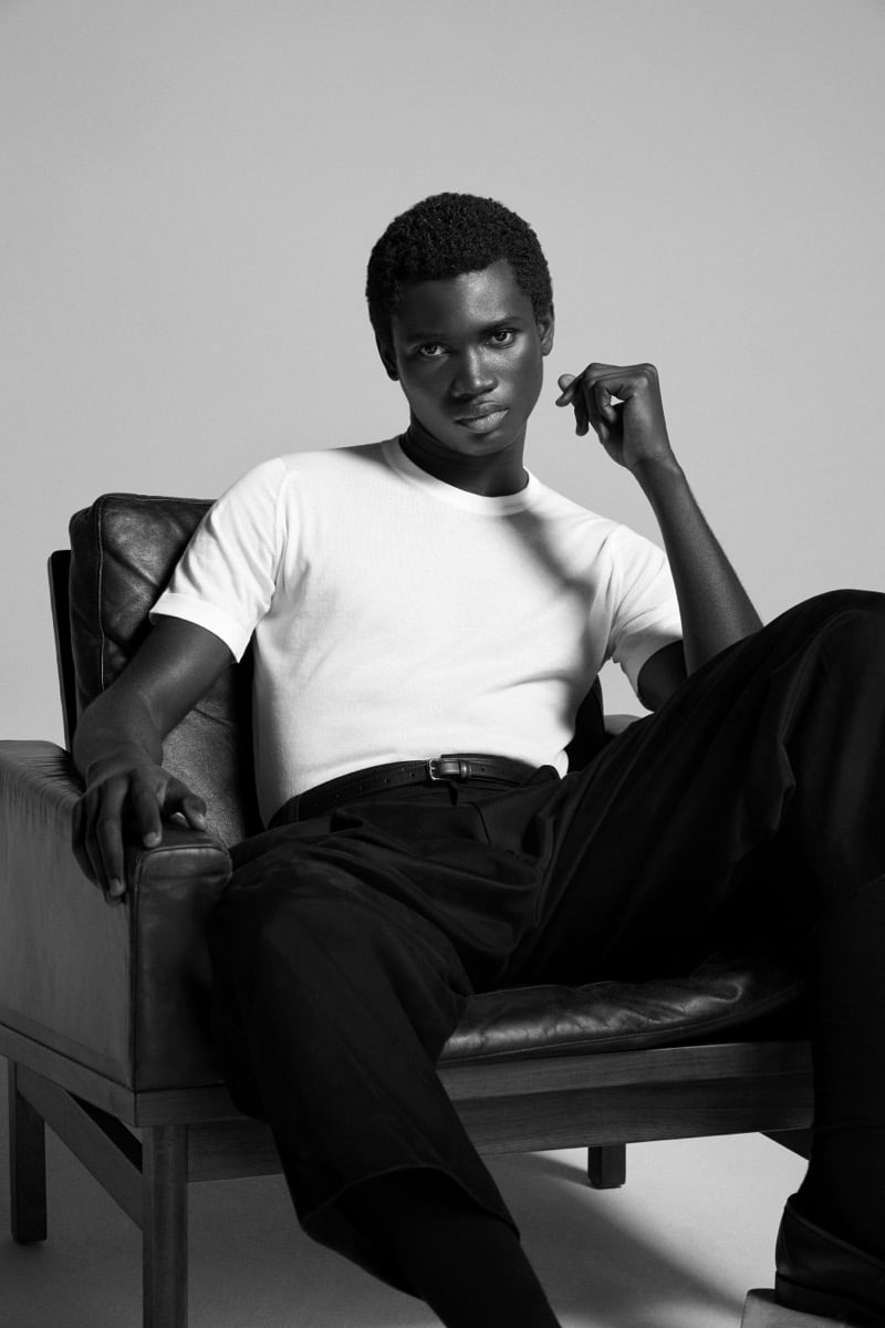 Alexander Acquah reclines in a leather chair wearing the John Smedley Torin Sea Island cotton T-shirt with dark pleated trousers and a slim leather belt