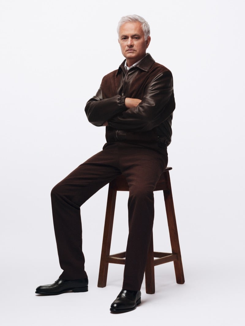 José Mourinho perched on a wooden stool with his arms crossed, wearing a chocolate brown leather bomber jacket over a white shirt with matching brown trousers and black leather loafers