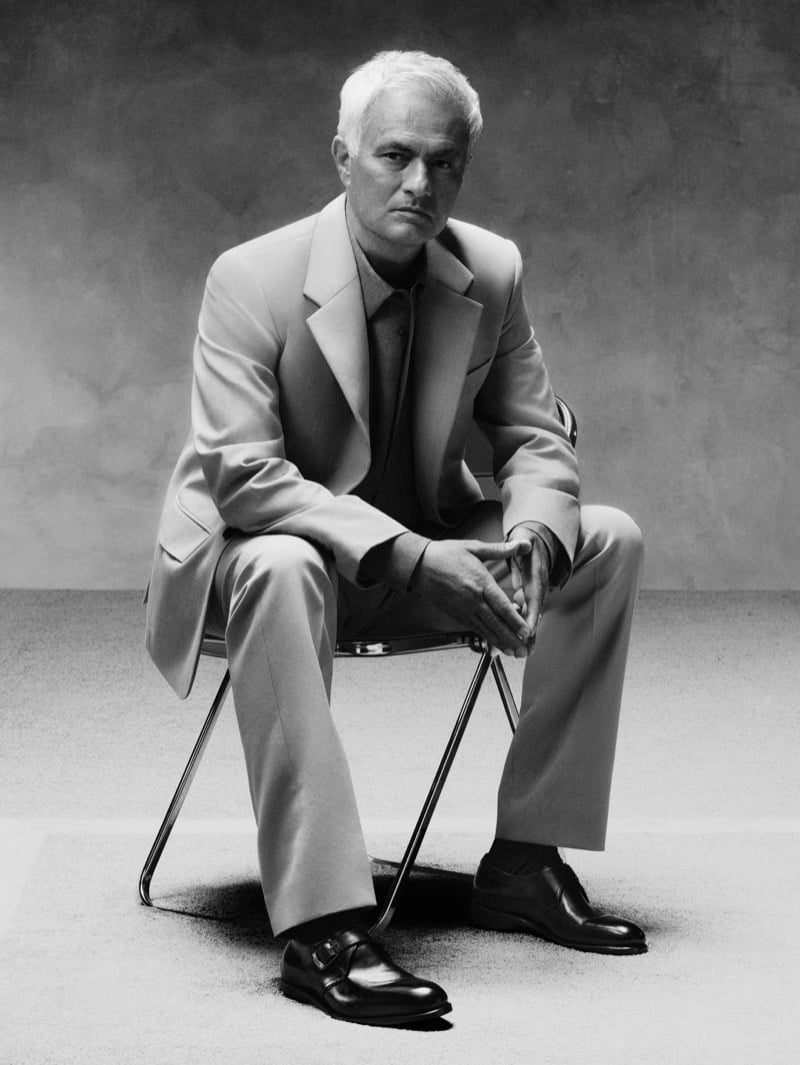 Black-and-white portrait of José Mourinho seated on a folding chair in a light tailored suit worn with a darker shirt and Ferragamo black leather monk-strap shoes