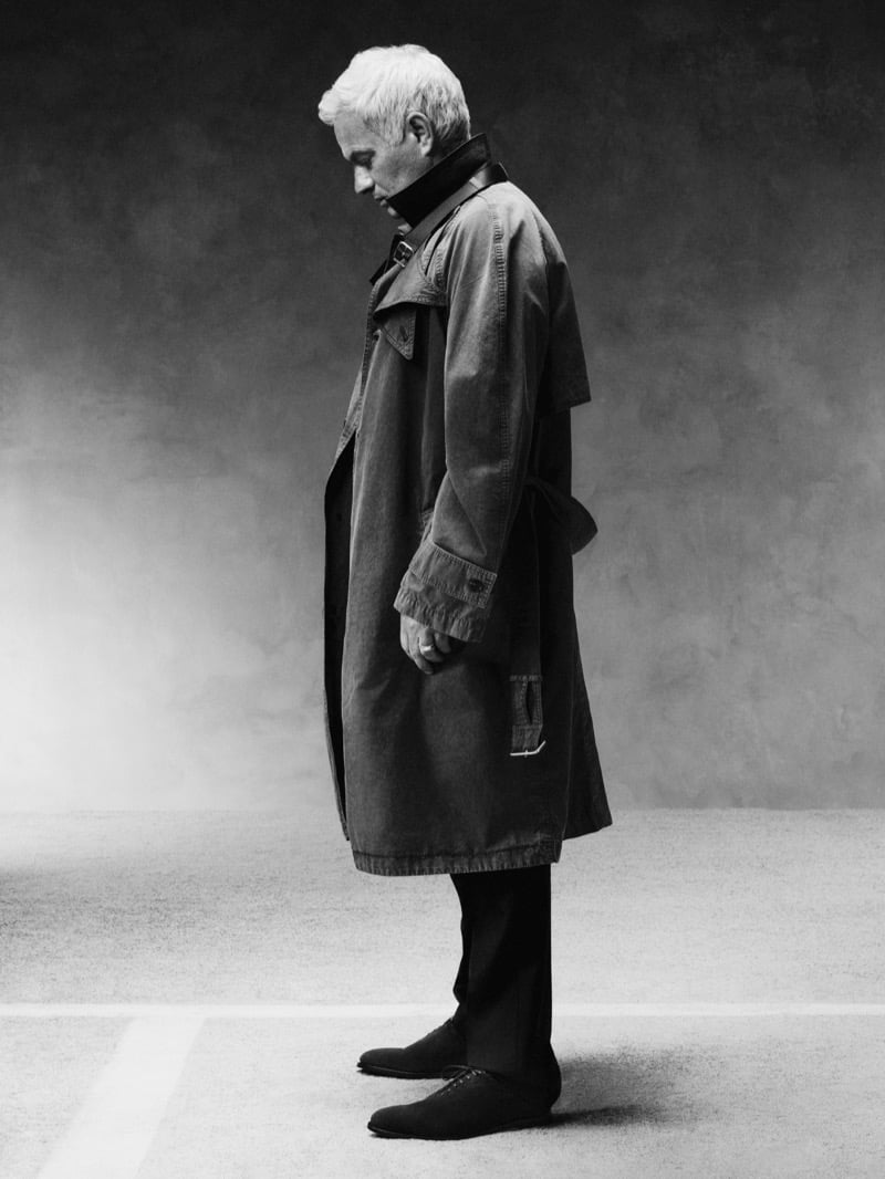 Black-and-white profile shot of José Mourinho standing with his head bowed in a cotton trench coat over dark trousers and dark lace-up shoes
