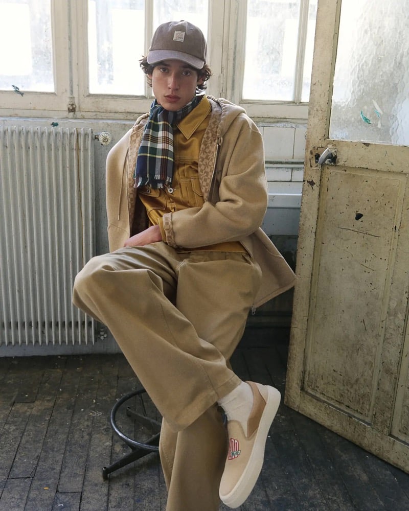 Max Fauviau sits on a stool in a brown corduroy Kenzo cap, plaid wool scarf, tan unstructured overcoat layered over a mustard chore jacket and plaid shirt, with tan trousers and tan slip-on sneakers