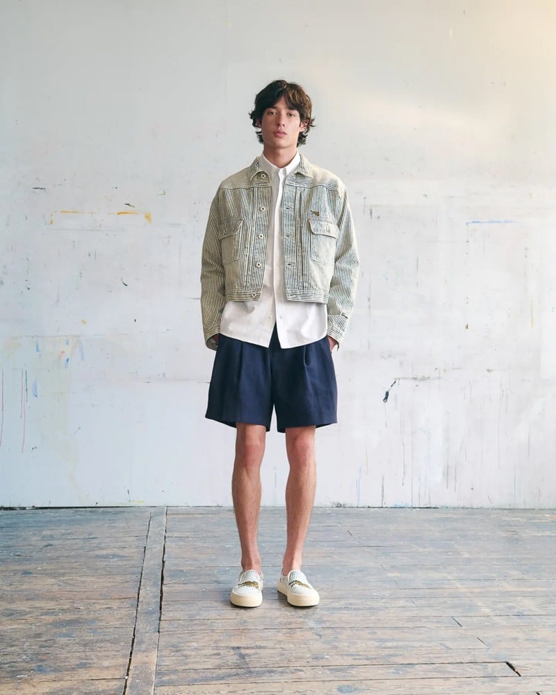 Max Fauviau in a cropped hickory-striped denim jacket over a white button-up shirt with pleated navy shorts and woven canvas slip-on sneakers against a white wall