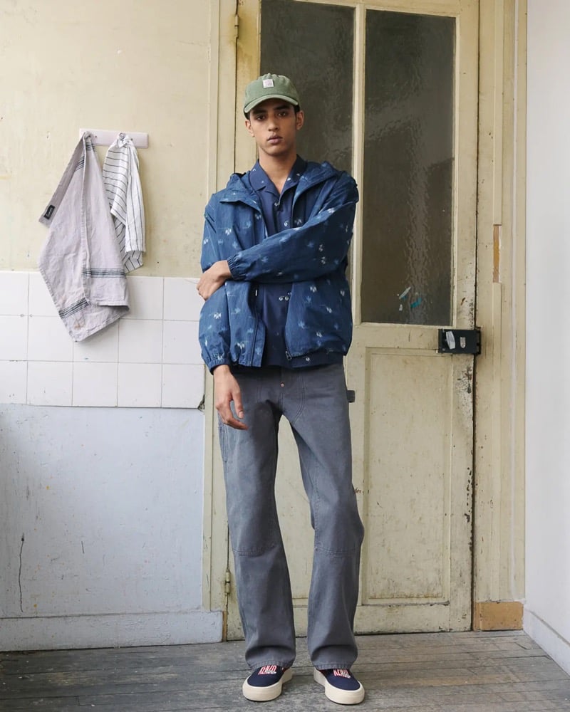 Abas Abdirazaq stands beside a kitchen door in an olive Kenzo cap, a blue floral-print hooded windbreaker over a navy camp shirt with gray carpenter trousers and Kenzo low-top sneakers