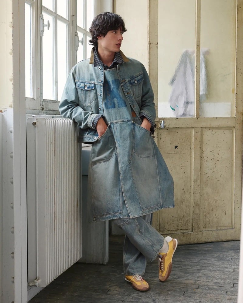 Max Fauviau leans against a radiator in a light wash denim trucker jacket layered over a long denim apron with a gingham shirt, blue jeans, and mustard and tan suede running sneakers
