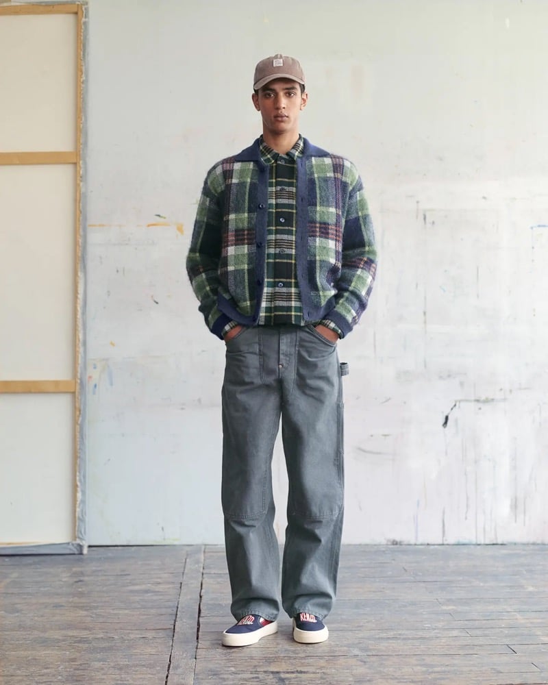 Abas Abdirazaq wears a brown corduroy Kenzo cap with a green plaid mohair cardigan over a matching green plaid flannel shirt, gray carpenter trousers, and navy and red Kenzo slip-on sneakers