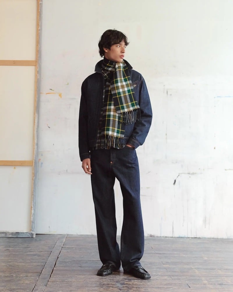 Max Fauviau stands in a raw indigo denim zip jacket with a green and yellow tartan fringe scarf, rinsed indigo jeans, and black tassel loafers