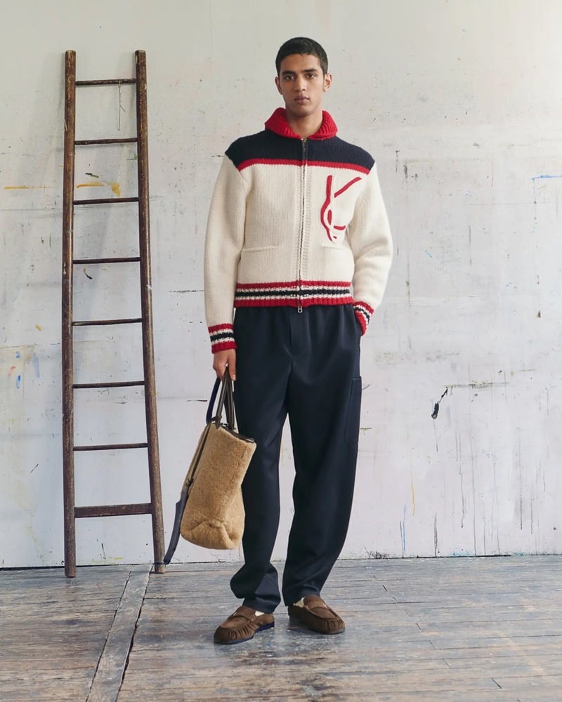 Abas Abdirazaq wears a cream cable-knit zip cardigan with a red collar and navy yoke featuring a red K graphic, paired with navy pleated trousers, brown suede moccasins, and a shearling tote bag