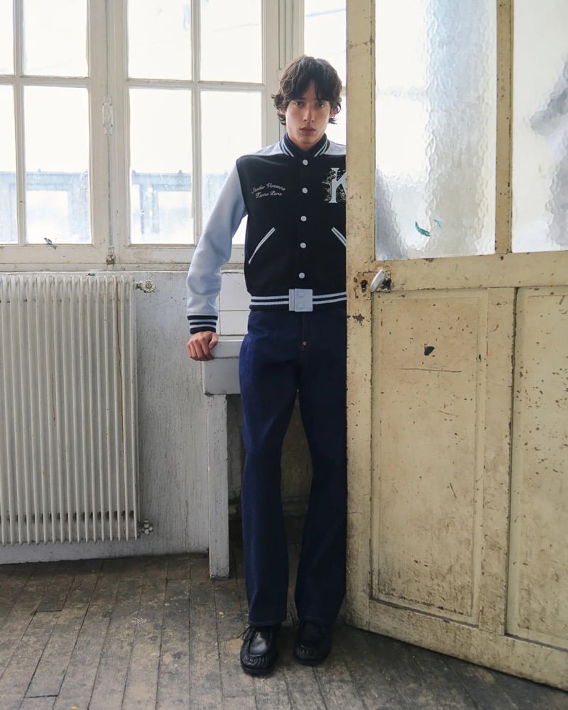Max Fauviau leans in a doorway wearing a black and gray Kenzo varsity jacket with embroidered K patch over dark rinse straight-leg jeans and black moccasin shoes