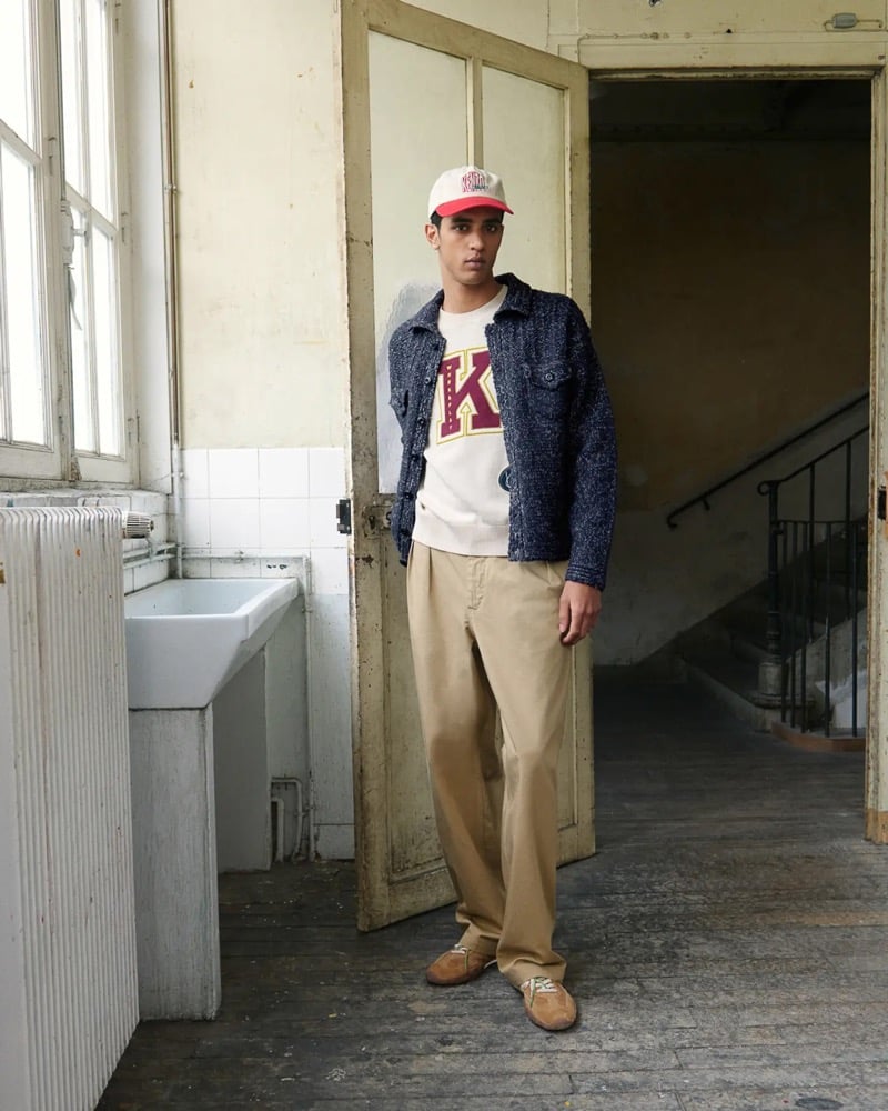Abas Abdirazaq stands in a doorway in a cream and red Kenzo varsity cap, a navy marled knit overshirt over a cream K-graphic sweatshirt, tan chinos, and brown suede sneakers