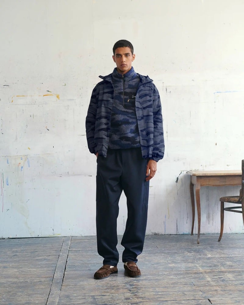 Abas Abdirazaq wears a navy camouflage-printed nylon zip jacket over a marled navy quarter-zip sweater with tapered navy cargo trousers and brown suede moccasins
