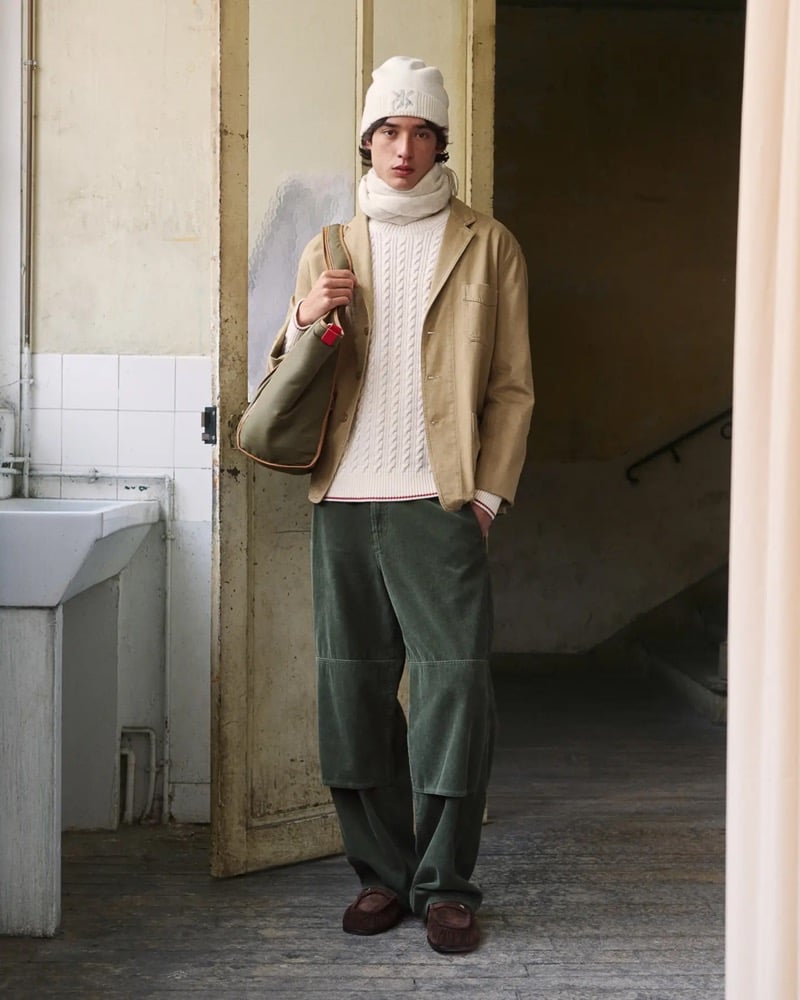 Max Fauviau stands in a doorway in a cream Kenzo beanie and matching cream cable-knit sweater layered under a tan unstructured blazer, with green corduroy trousers and brown suede moccasins, an olive tote bag at his shoulder