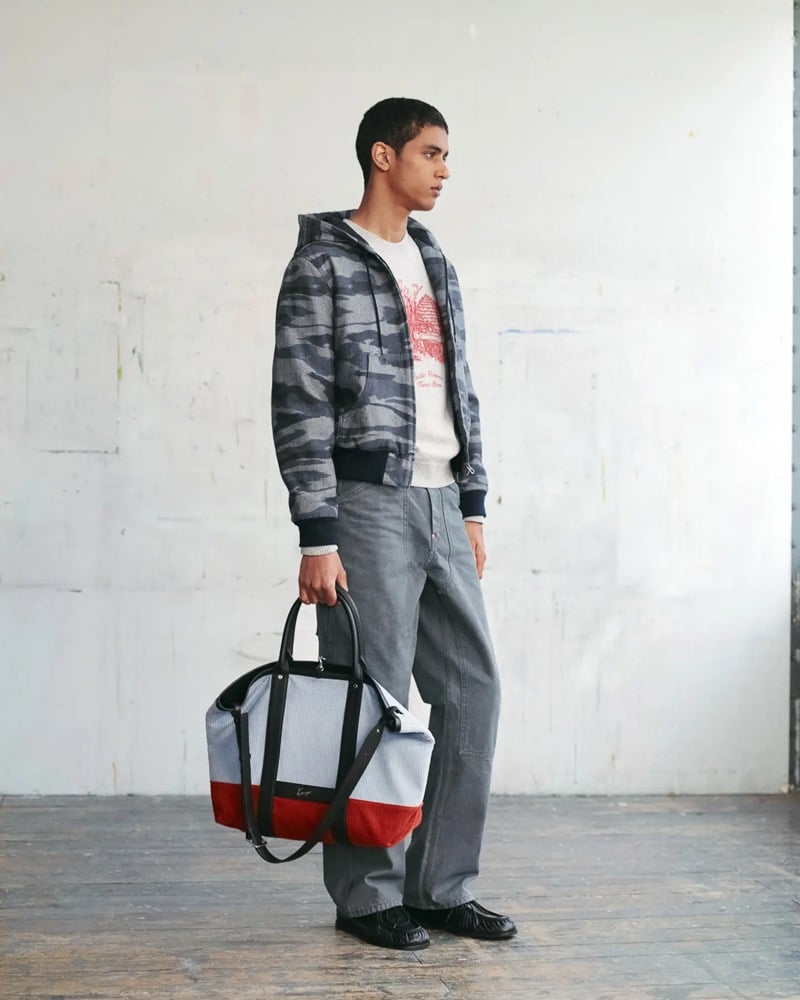 Abas Abdirazaq wears a gray camouflage hooded sweatshirt over a cream crewneck printed with a red Kenzo eagle graphic, paired with gray carpenter trousers, a striped Kenzo tote, and black loafers