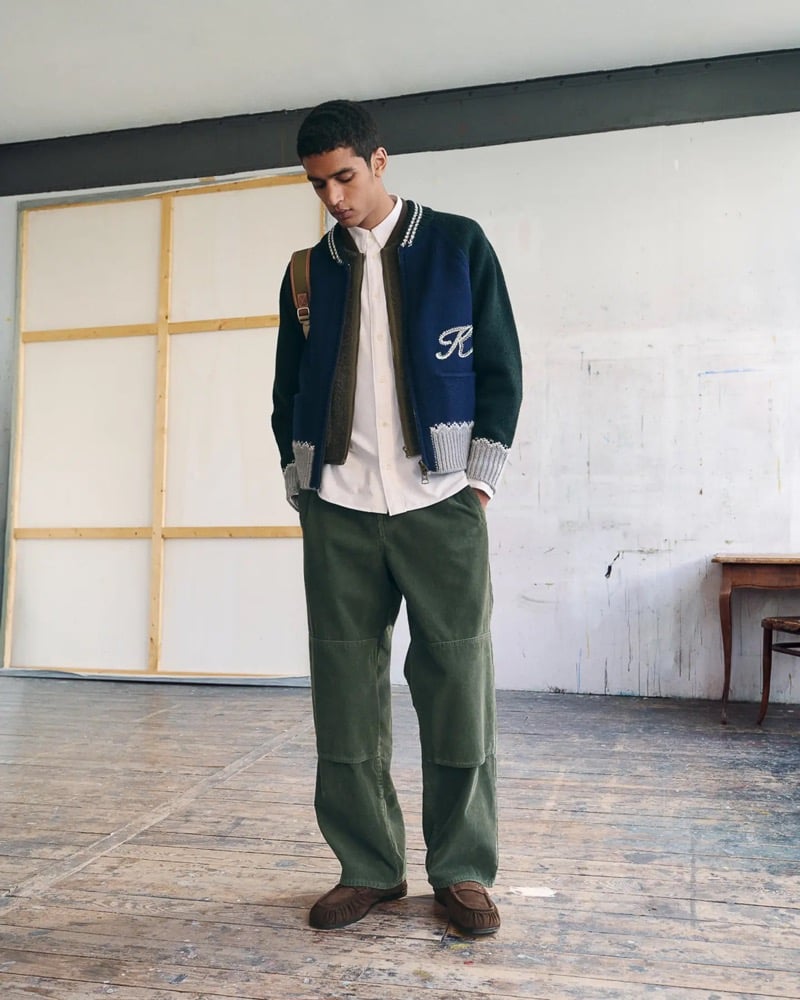 Abas Abdirazaq wears a navy and forest green Kenzo varsity sweater jacket with white K script and Fair Isle trim over a white shirt, olive carpenter trousers, and brown suede moccasins