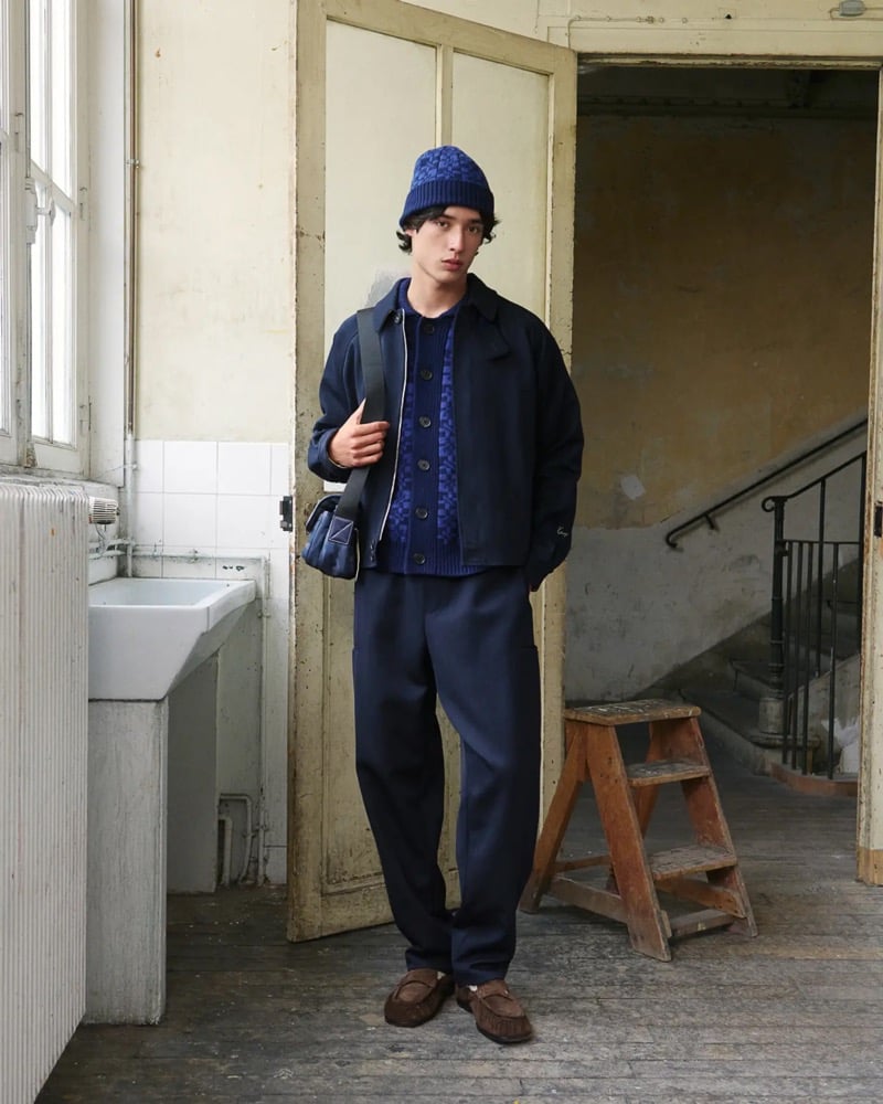 Max Fauviau stands in a doorway in a navy textured beanie, a navy zip overshirt layered over a checkered navy knit cardigan, navy pleated trousers, and brown suede moccasins, a small navy crossbody bag at his hip