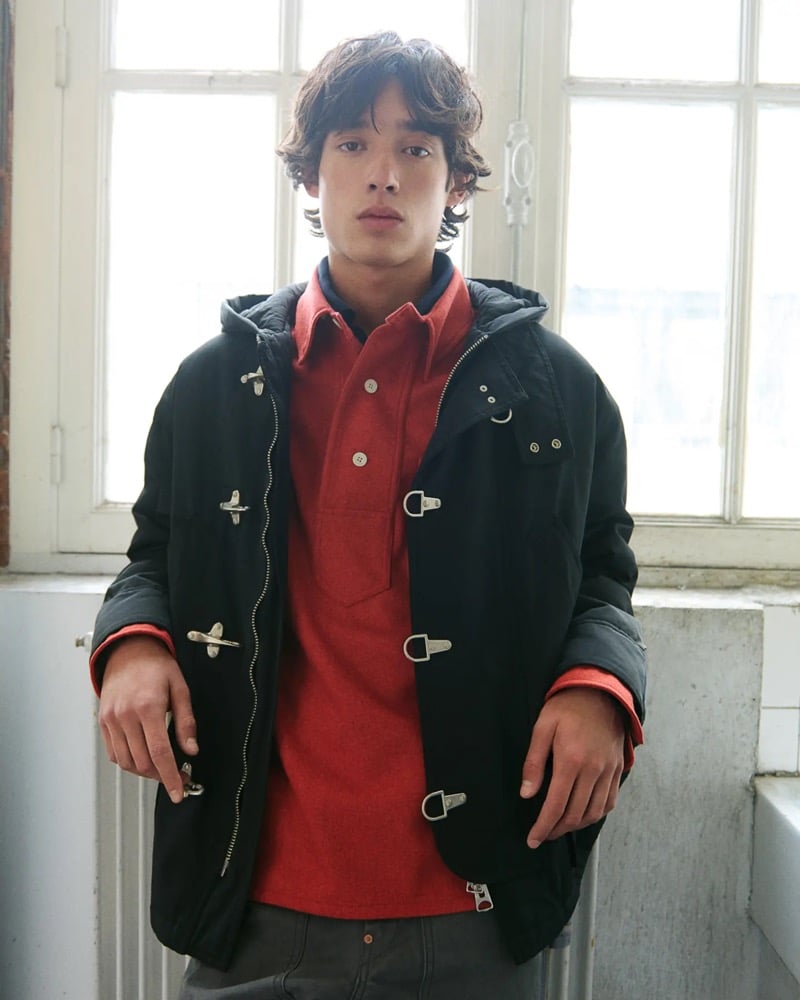 Close-up of Max Fauviau in a black hooded duffle parka with silver toggle clasps over a red collared rugby polo and gray carpenter jeans