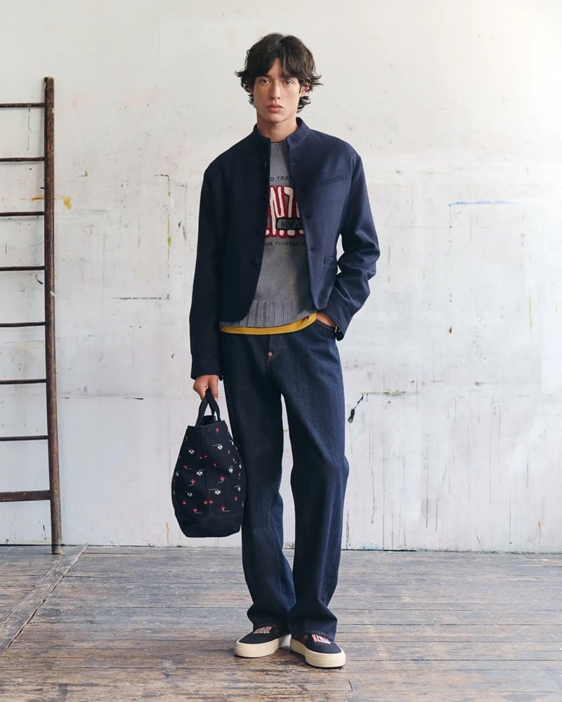 Max Fauviau wears a navy collarless mandarin blazer over a gray varsity sweater printed with a 1970 Rue Vivienne graphic, dark rinse jeans, and navy Kenzo slip-on sneakers, holding a floral-printed canvas tote