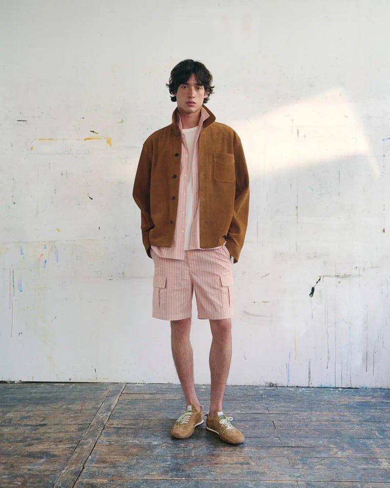 Max Fauviau wears a tan suede chore jacket over a pink striped seersucker shirt and white tee with matching pink striped cargo shorts and tan and green suede running sneakers