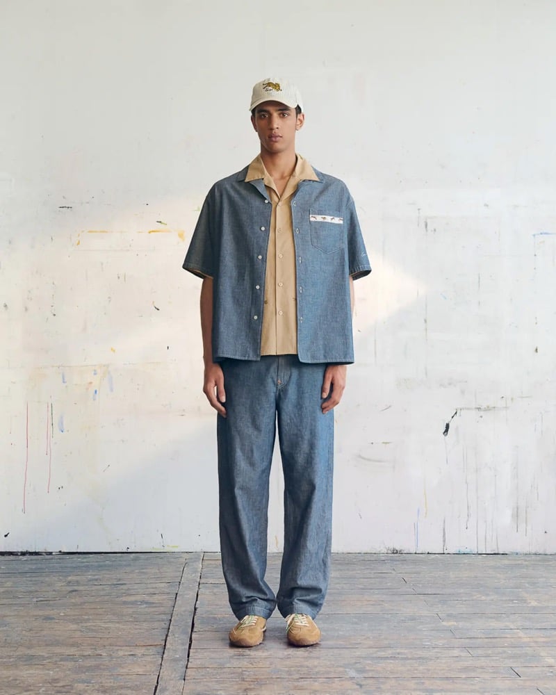 Abas Abdirazaq wears a cream corduroy Kenzo tiger-embroidered cap, a chambray short-sleeve camp shirt layered over a tan camp shirt, chambray jeans, and tan suede sneakers