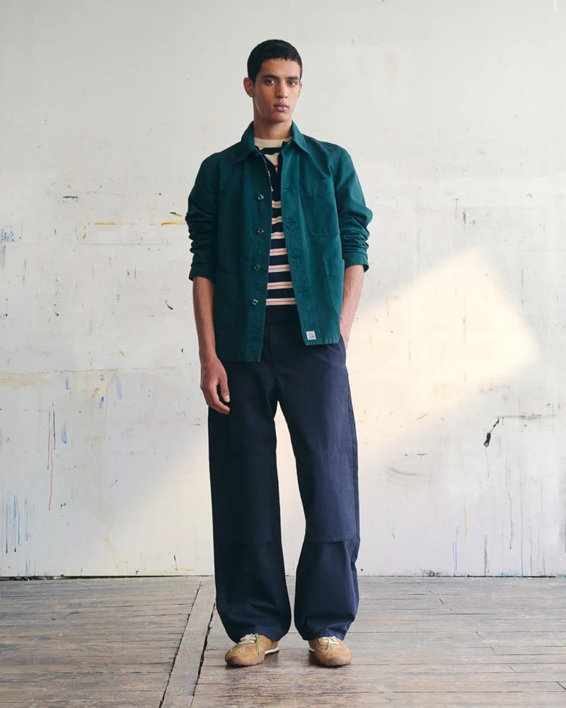 Abas Abdirazaq wears a teal green French chore jacket over a navy and peach striped knit, navy carpenter trousers, and tan suede running sneakers