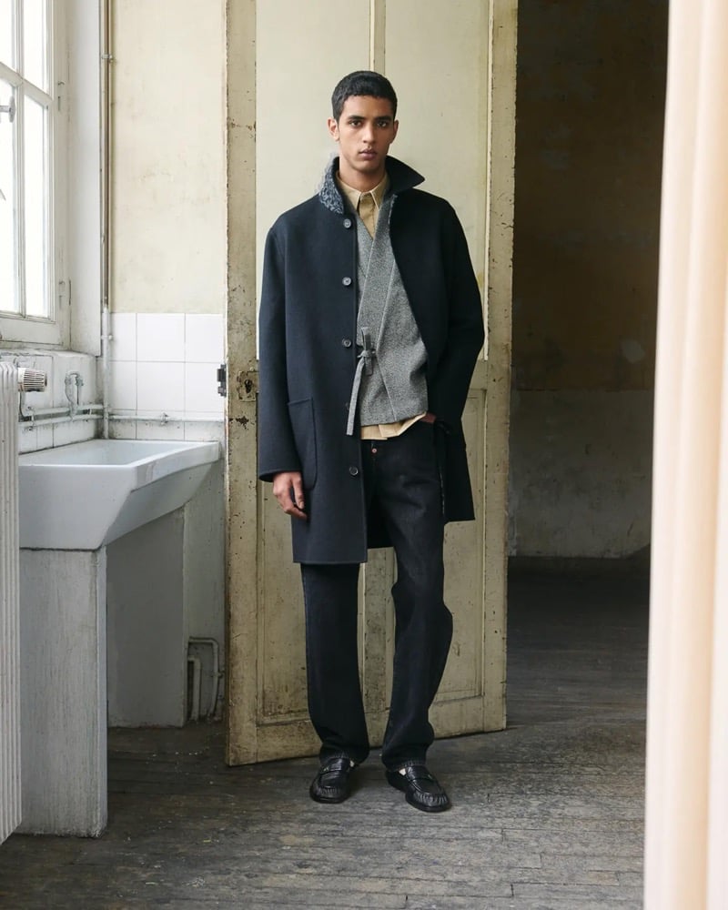 Abas Abdirazaq stands in a doorway in a dark navy wool single-breasted coat layered over a gray cable cardigan with a mustard polo, dark wash jeans, and black tassel loafers