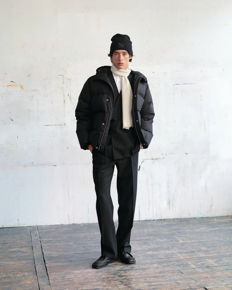 Max Fauviau wears a black Kenzo K beanie, a black down puffer jacket with quilted floral panels over a black pinstripe suit, a cream wool scarf, and black tassel loafers