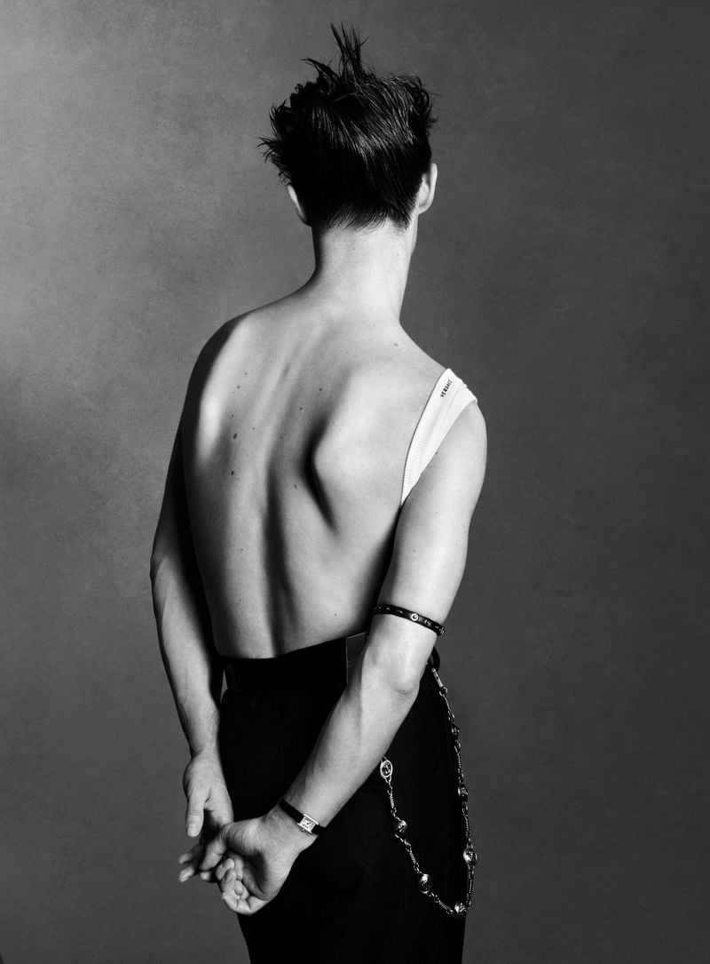 Kit Butler photographed from behind, bare back, one arm threaded through the strap of a white Versace bodysuit, low-rise black trousers with a long silver chain swinging at the hip, hands clasped at the small of his back, watch and studded leather bracelet on his wrist