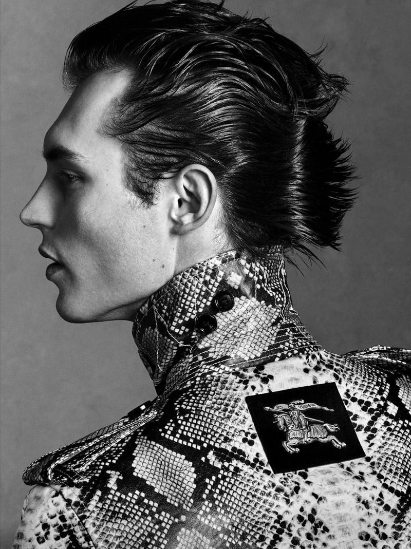 Kit Butler in close profile portrait wearing a Burberry python-print high-neck coat buttoned tight at the neck, wet swept-back hair against a neutral gray background