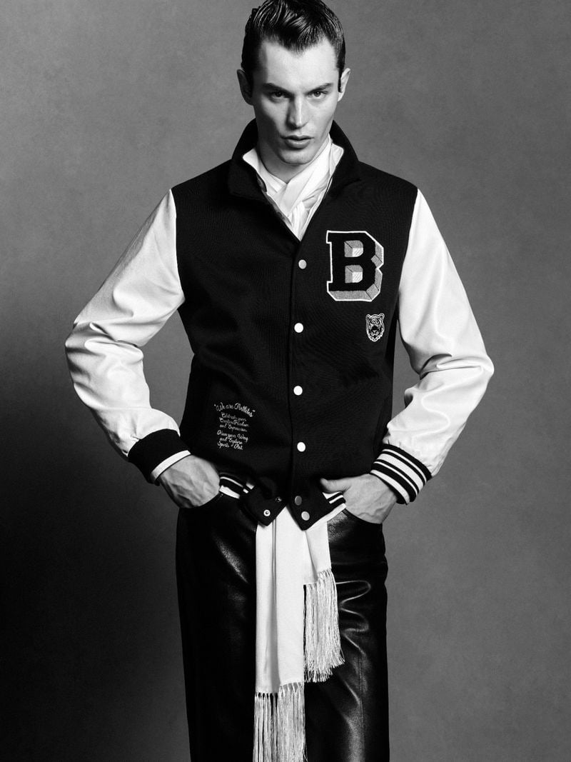 Kit Butler in a Bench varsity letterman jacket with a black wool body, white leather sleeves, chenille B letter and tiger emblem, layered over a fringed white silk scarf trailing down, paired with a black leather pants, hands in pockets