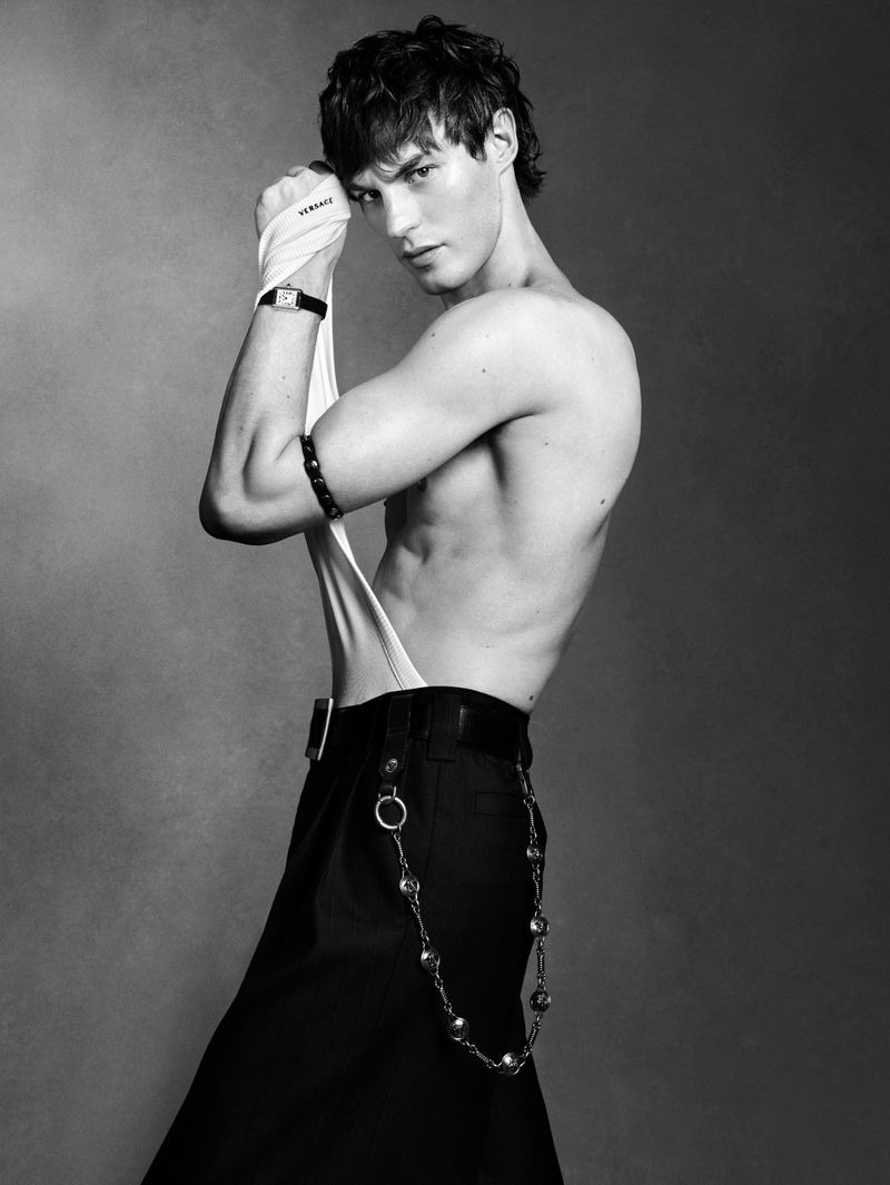 Kit Butler bare-chested in three-quarter profile, lifting a white Versace bodysuit toward his head, a square tank-style wristwatch and studded leather bracelet on his arm, low-rise black pinstripe trousers with a long silver medallion chain swinging from the belt loops