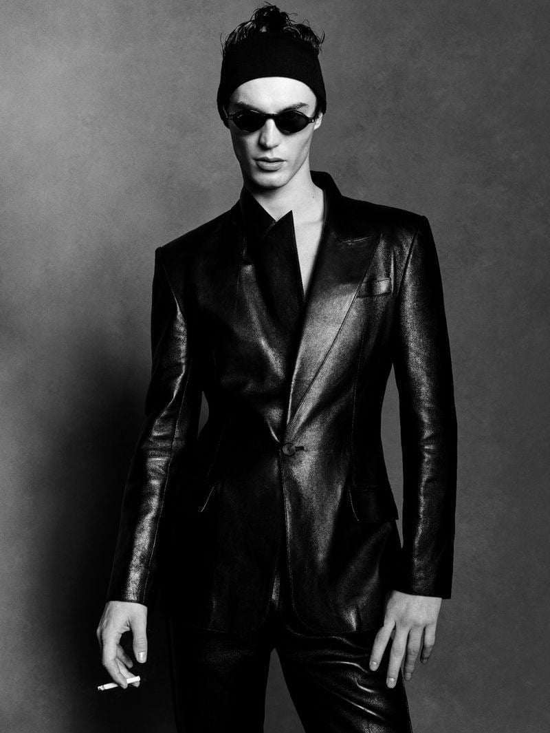 Kit Butler in a slim black leather suit with a sharp peak-lapel single-button blazer and matching narrow trousers, narrow oval black sunglasses, a lit cigarette held in his right hand