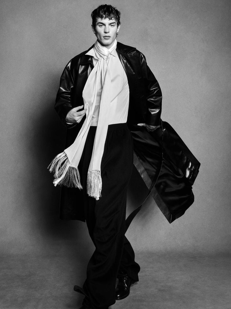 Kit Butler walking forward in a black leather midi trench coat swinging open at the side, a long white silk fringed scarf wrapped at the neck trailing down over a white shirt and black wide-leg trousers, finished with black leather boots