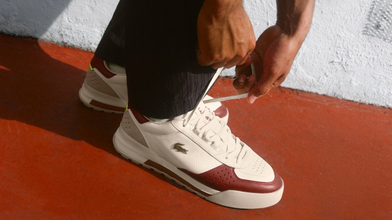 On-foot view of Arthur Fils tying the laces of the Lacoste Slam Break Premium sneaker in off-white leather with deep red side panels, a neon green pull tab at the heel, and a metal crocodile logo, worn over dark crinkle shorts with white socks against a clay-red painted ground beside a whitewashed wall
