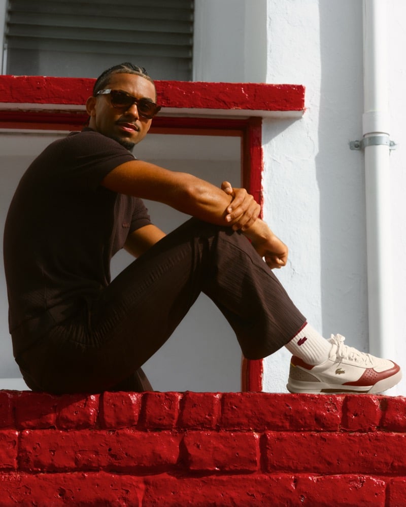 Arthur Fils seated on a glossy red-painted brick ledge in dark sunglasses, a dark brown short-sleeve piqué polo, and dark crinkle wide-leg trousers, the white and red Slam Break Premium sneaker on his foot with a white crocodile-logo sock, framed by a window trimmed in matching red