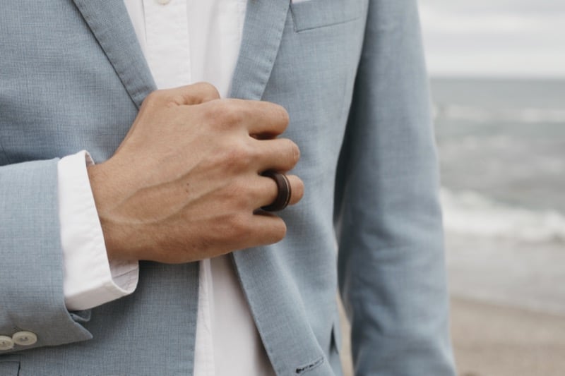 Cropped shot of a man at the shoreline in a dusty blue linen suit jacket, his hand adjusting the front near the chest with a small pocket square visible, the slubby flax weave catching afternoon light