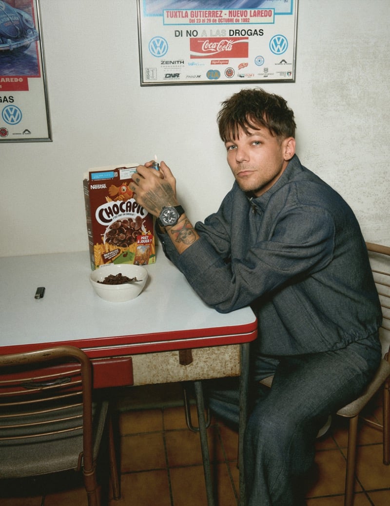 Louis Tomlinson sits at a red-trimmed retro diner table with a box of Chocapic cereal and a small bowl in front of him, wearing a Fendi denim shirt jacket and matching pants with a Hublot watch on his wrist