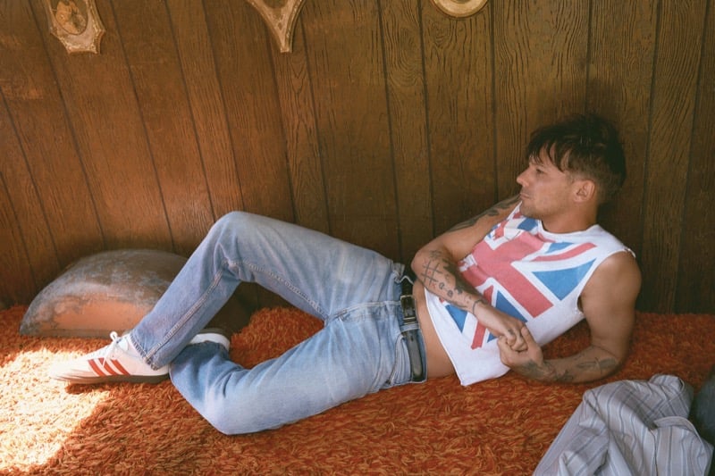 Louis Tomlinson reclines on an orange shag rug in a vintage Union Jack flag tank top, light wash Levi's jeans, and white Adidas Originals sneakers with red stripes