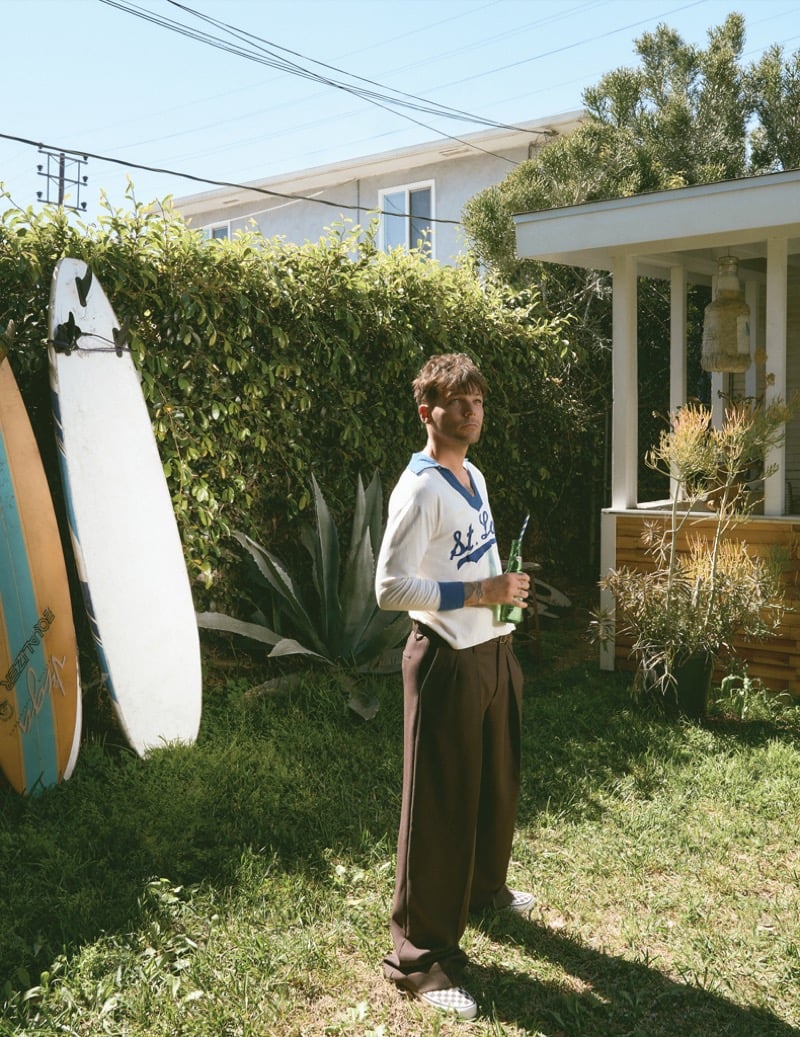 Louis Tomlinson stands in a sunlit backyard beside a row of surfboards wearing a vintage striped collared knit pullover and wide pleated brown trousers with checkerboard Vans slip-ons
