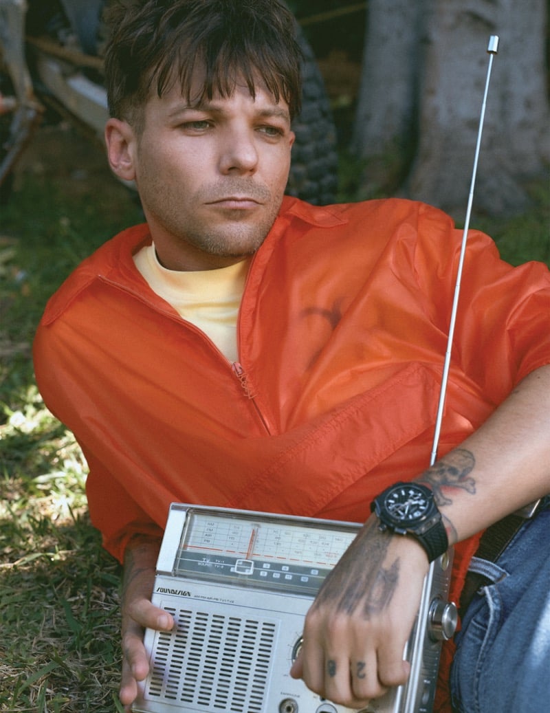 Louis Tomlinson sits on the grass in an open orange Saint Laurent zip windbreaker over a pale yellow tee and Agolde jeans, holding a vintage Soundesign radio with a Hublot watch on his wrist