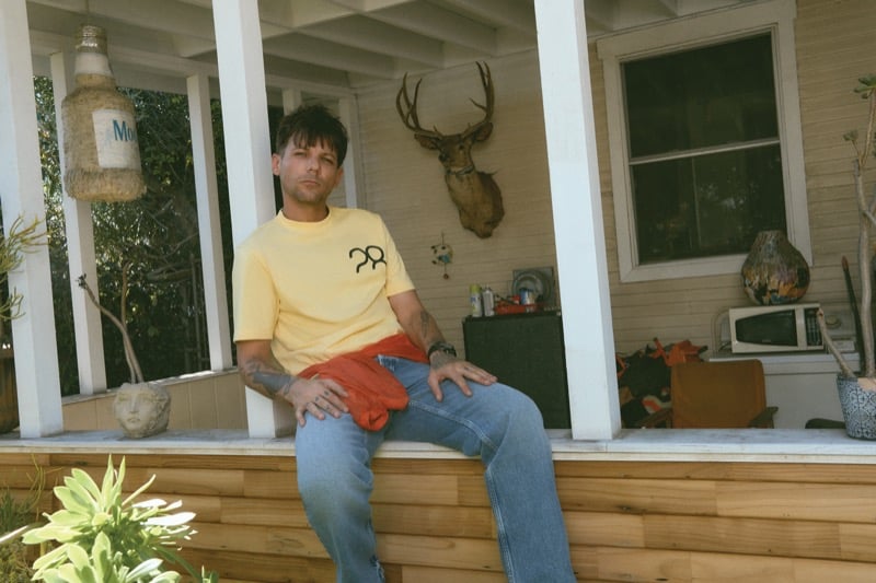 Louis Tomlinson sits on a porch ledge in a pale yellow logo tee with an orange Saint Laurent jacket tied at the waist over relaxed Agolde jeans, accessorized with a Hublot watch