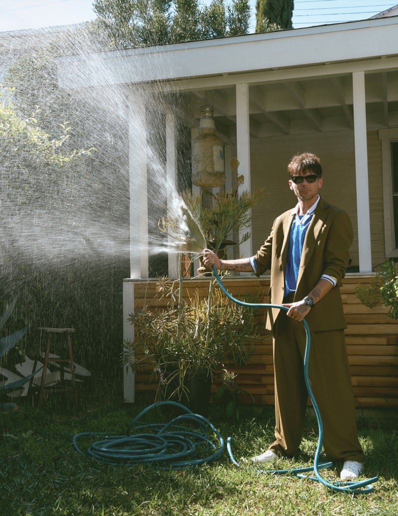 Louis Tomlinson holds a garden hose in front of a porch wearing a tobacco brown Tod's suit over a bright blue striped jersey, paired with Tretorn sneakers and Oliver Peoples sunglasses