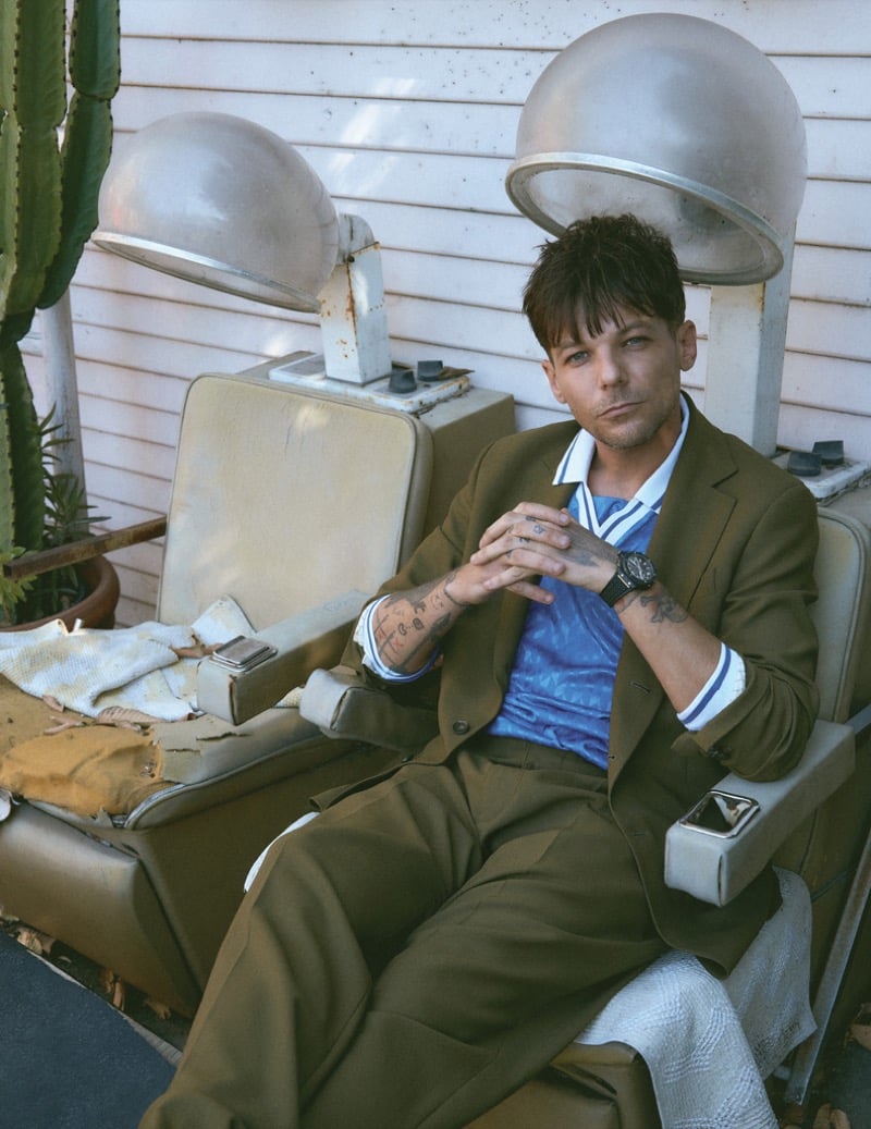 Louis Tomlinson reclines in a vintage salon dryer chair in a tobacco brown Tod's suit over a bright blue striped jersey