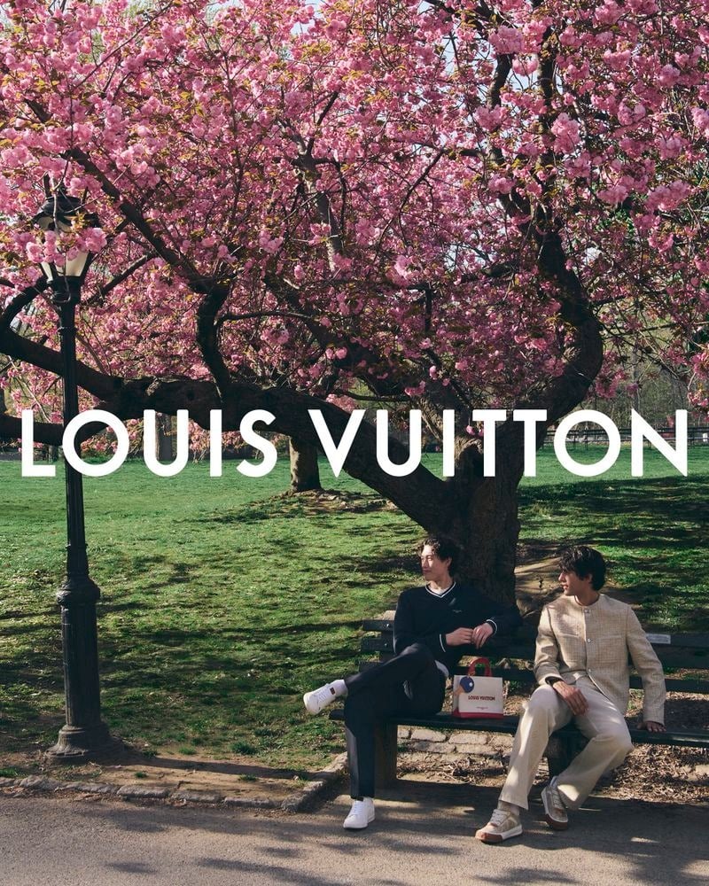 Mathieu Simoneau and Akbar Shamji sit on a Central Park bench beneath a blooming pink cherry blossom tree, with Simoneau in a navy track set and white sneakers and Shamji in cream linen trousers with a white tee